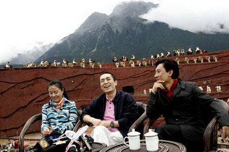 Director Zhang Yimou (C) answers questions as actors dressed in ethnic costumes perform behind him in an outdoor production called 